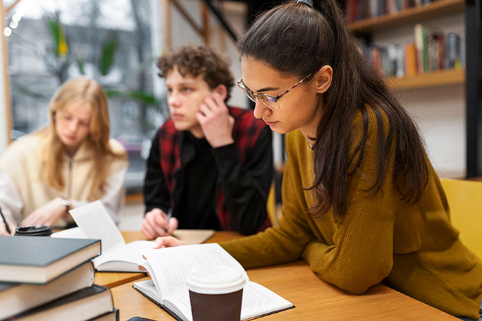 Young woman focused on reading a book with intense intelligence surrounded by classmates in a library setting.