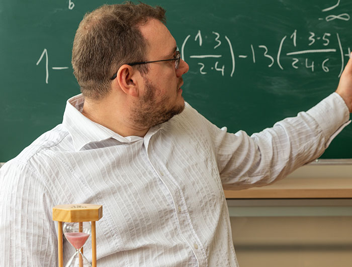 Man wearing glasses and white shirt writing complex math formulas on chalkboard demonstrating sharp intelligence.