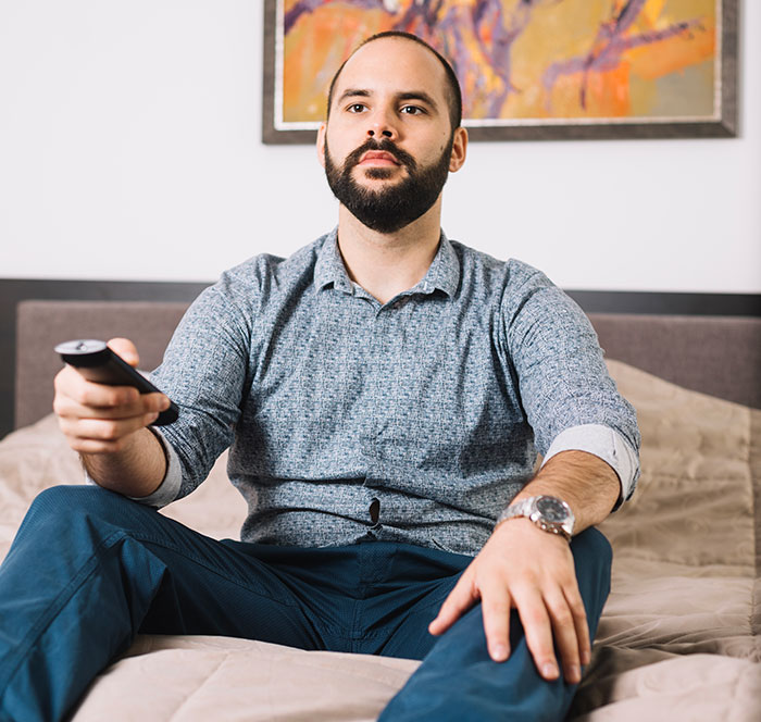 Man with beard sitting on bed holding remote control, focused expression showing sharp intelligence and deep thought.