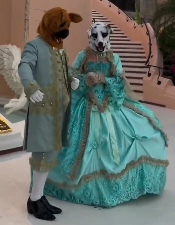 Two people in dog masks and old-time costumes pose at a Mar-a-Lago event with a dramatic staircase backdrop.