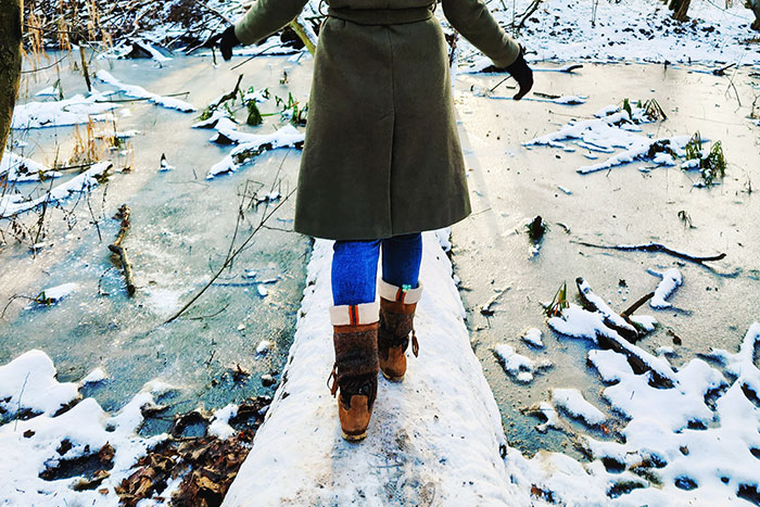 Person wearing brown boots and a coat carefully walking on a snowy log over frozen water, showcasing dangerous things people did.