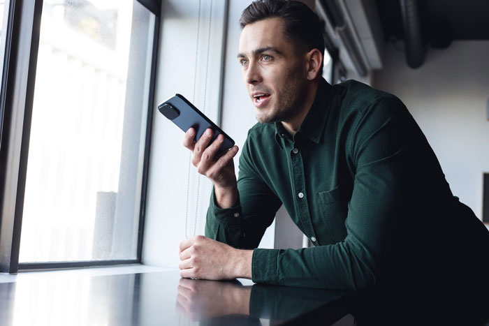 Man holding smartphone near window, appearing surprised while talking inside a modern cafe or bar setting. Man holding smartphone near window, appearing surprised while talking inside a modern cafe or bar setting.