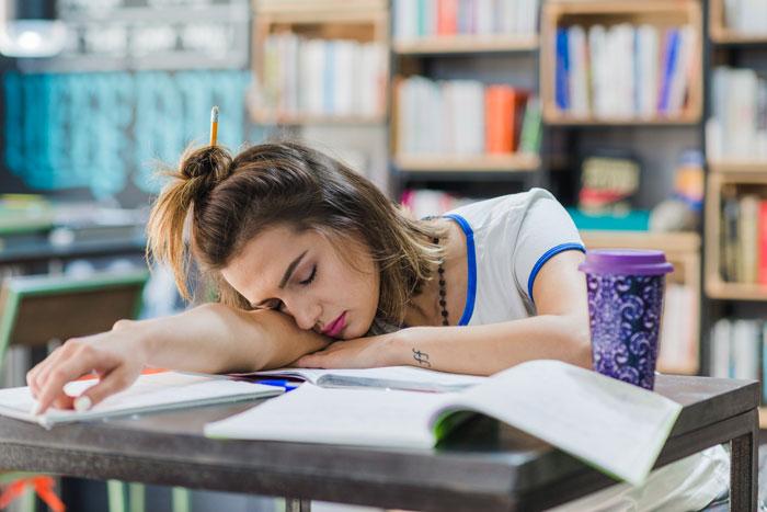 Young daughter resting head on arm at table, surrounded by open books and a drink, ready to step up and help mom.
