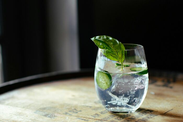 Glass of sparkling water with cucumber slices and basil leaf on a rustic wooden surface, no shipping containers visible.