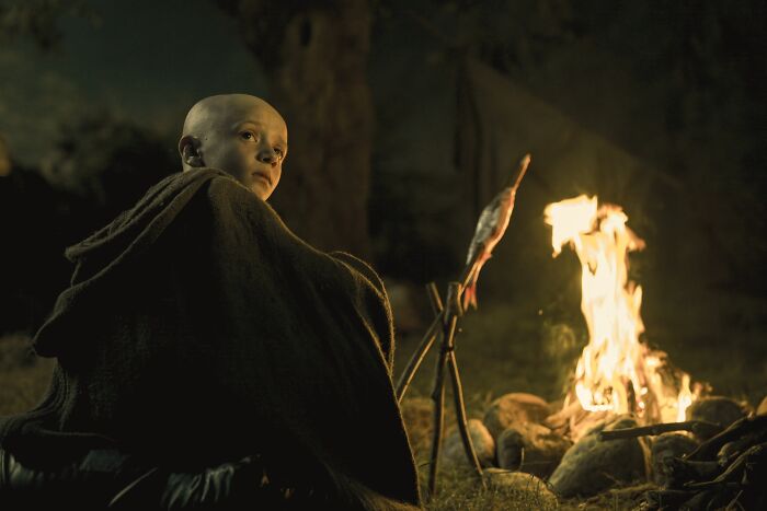 Bald character wrapped in a cloak sitting by a campfire roasting fish, related to Egg from A Knight of the Seven Kingdoms Game of Thrones.