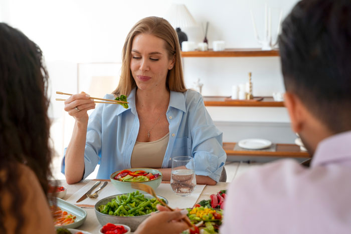 Woman eating at a family dinner while others, including a man, sit around the table in a bright room. Woman eating at a family dinner while others, including a man, sit around the table in a bright room.
