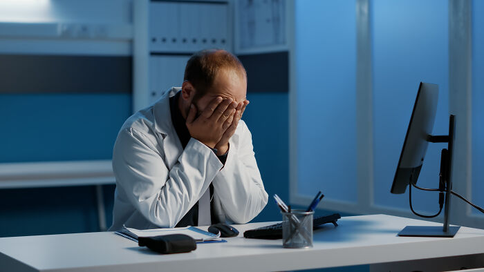 Doctor in white coat sitting at desk covering face with hands, stressed after encounter with anti-vaxxer in medical office.
