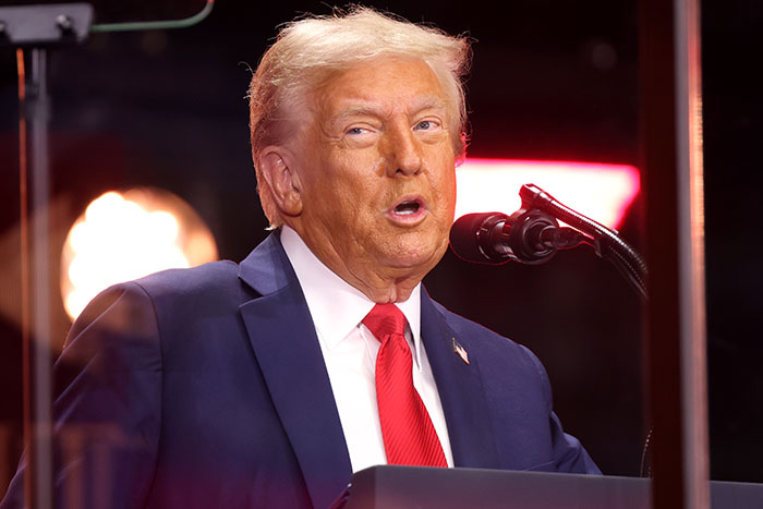 Former President Donald Trump speaking at a podium in a suit and red tie during a public event.