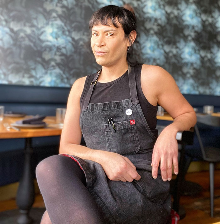 Trans chef of the year sitting in a restaurant wearing a black apron and sleeveless shirt, posing confidently. Trans chef of the year sitting in a restaurant wearing a black apron and sleeveless shirt, posing confidently.