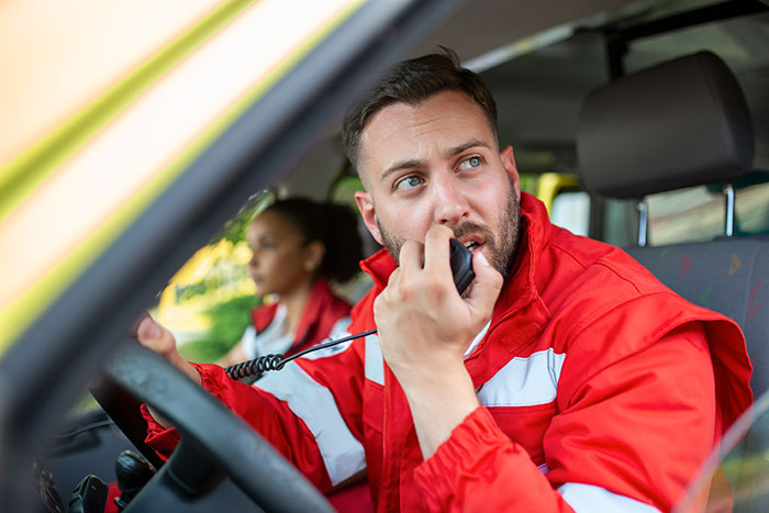 Male paramedic in red uniform speaking into radio in ambulance, colleague in background, conveying info while calling 911