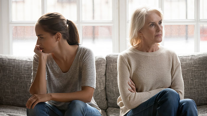 Middle-aged woman and younger woman sitting apart on couch looking upset, showing tension and mistreatment issues.
