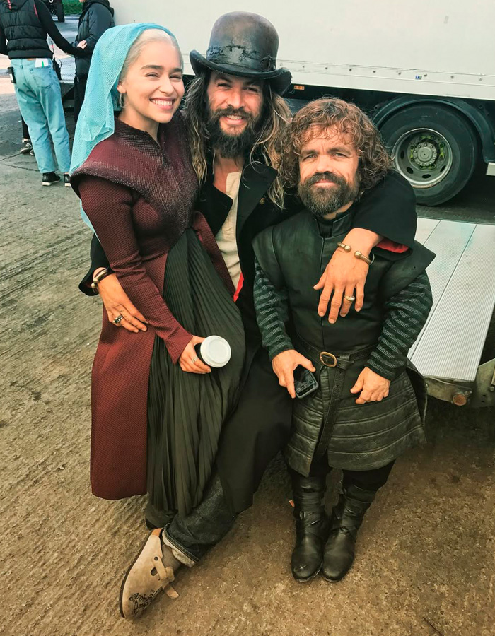 Emilia Clarke smiling with two men in costume on set, relating to her broken rib during intimate scene filming.
