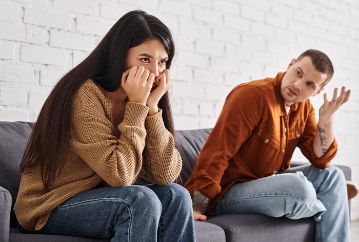 Woman upset on couch threatening to take ex&rsquo;s dog to shelter while ex looks on, not intervening in the argument.