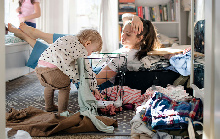 Tired woman lying on sofa while child plays with laundry in a cluttered living room setting.