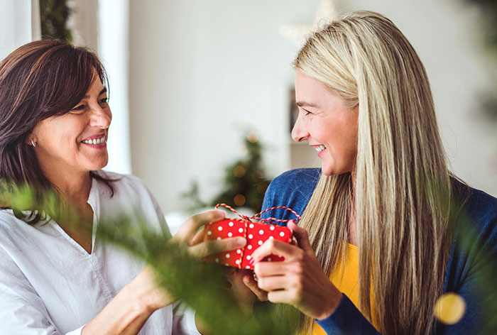 Two women smiling and exchanging a Christmas gift, illustrating a woman discovering her engagement to her first cousin.