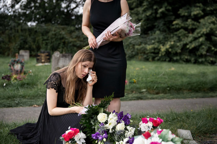 Two women in black mourning at a gravesite, one holding flowers, the other crying, reflecting engagement hold late boyfriends ashes.