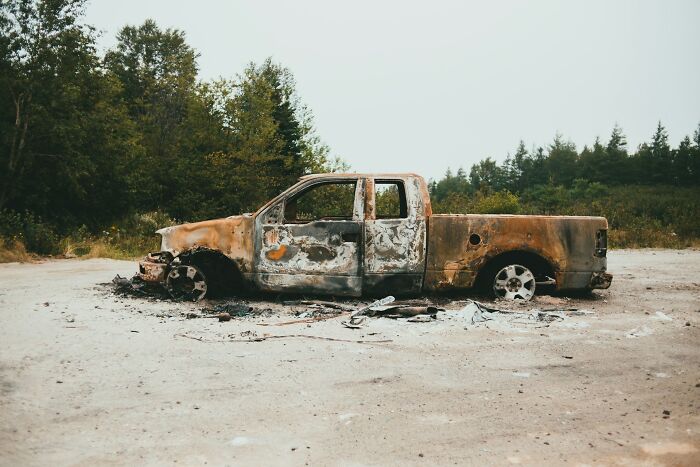 Burned-out and abandoned pickup truck on a dirt road surrounded by trees, showing shocking public destruction without shame.