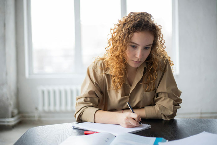 Young woman with curly hair focused on writing notes at a table, reflecting on ex house drama relationship issues.