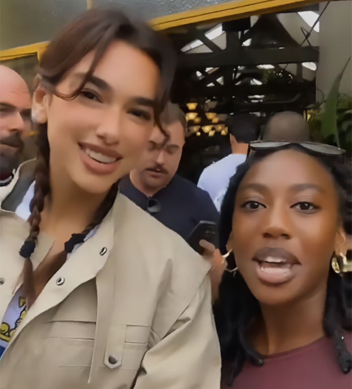 Dua Lipa smiling with a fan using a filter on their selfie, captured in a casual indoor setting with people in the background
