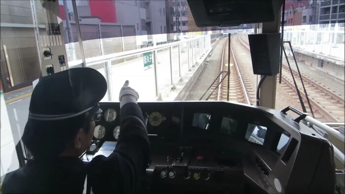 Train operator in uniform inside the cabin pointing ahead with controls and railway tracks visible, illustrating transport logistics challenges.