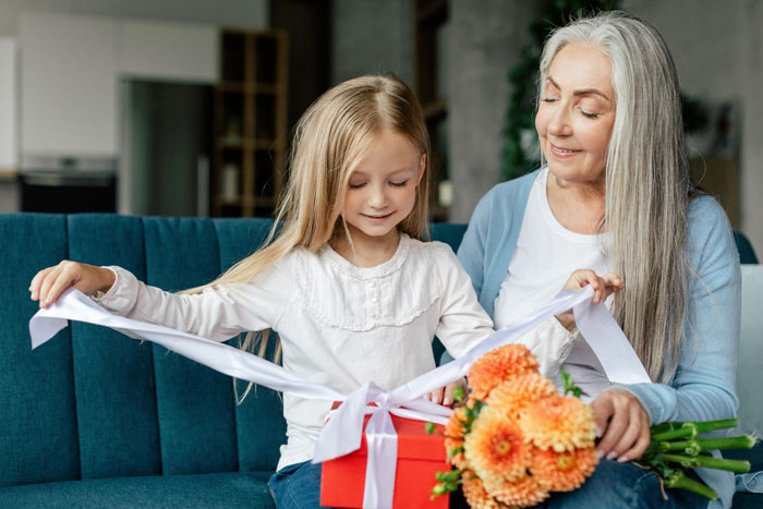Grandmother and granddaughter on a couch, granddaughter opening a gift box, highlighting favoritism in family celebrations.