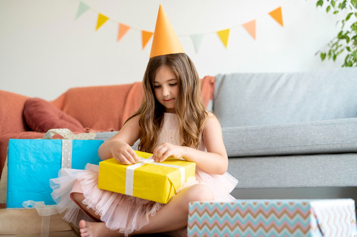 Young girl in a party hat opening a gift, highlighting aunties' favoritism and mom's frustration over one kid.