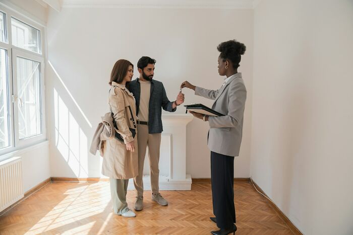 Real estate agent handing house keys to young couple in empty room, symbolizing adult goals and milestones.