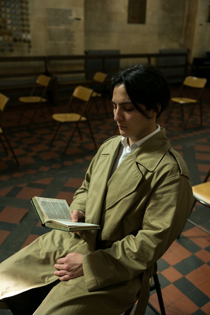 Young man in a beige trench coat sitting alone reading a book in an empty room, reflecting on cult member experiences.
