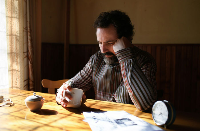 Man sitting at wooden table with cup, looking troubled and reflecting on his children as failures in his eyes.