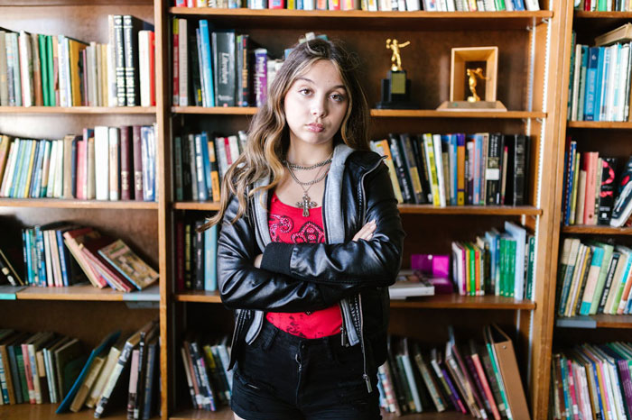 Teen girl with crossed arms standing in front of a bookshelf, reflecting on dad's claim of children being failures.
