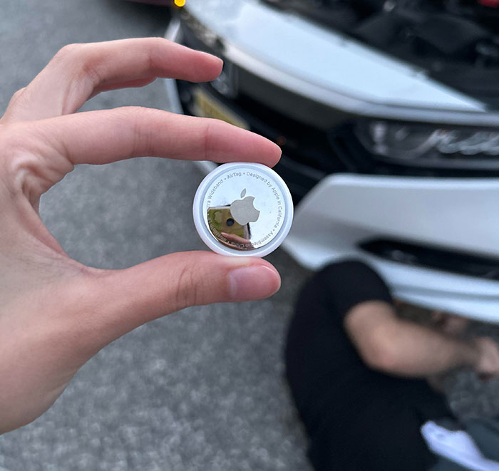 Hand holding an Apple AirTag in front of a white car with a person lying on the ground near the vehicle.