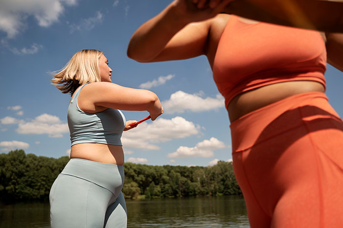 Two women in athletic wear exercising outdoors near a lake, highlighting women's bodies and fitness.