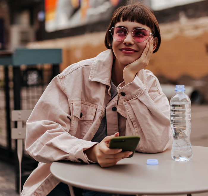 Young woman wearing sunglasses, sitting at outdoor cafe table with phone and water bottle, reflecting on women’s bodies.