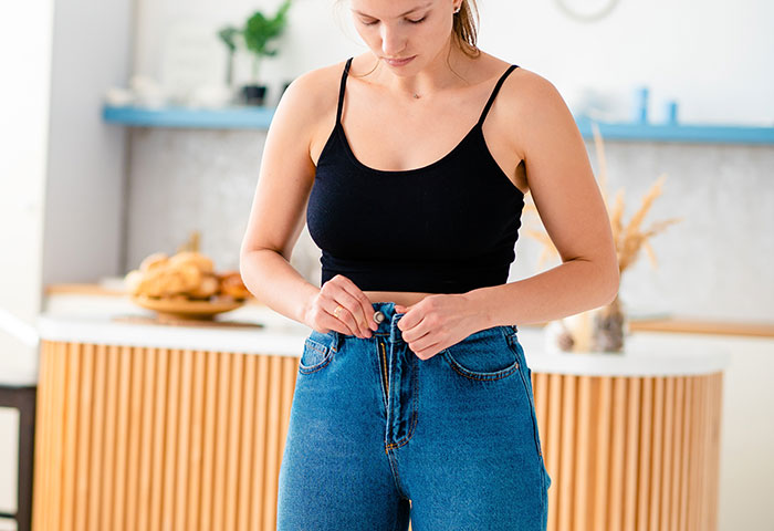 Young woman wearing a black tank top and blue jeans, focusing on buttoning jeans, highlighting women's body awareness.