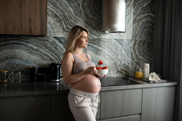 Pregnant woman eating fruit in modern kitchen, reflecting on women's bodies and health knowledge earlier on.