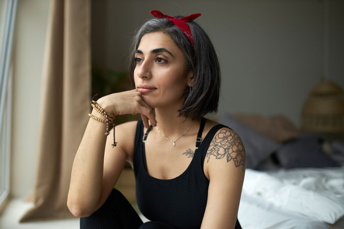 Woman with floral tattoo on shoulder wearing a red headband, looking thoughtful indoors by a window in casual wear.