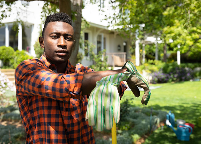 Young man in a plaid shirt wearing gardening gloves in front of a suburban house, illustrating homeownership challenges.