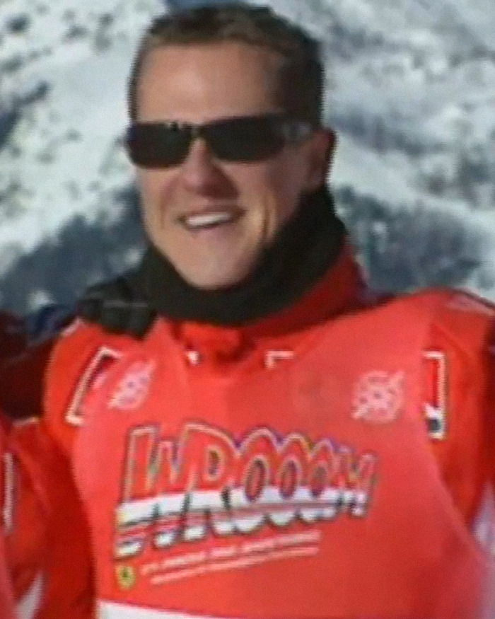 Michael Schumacher smiling outdoors in a red ski jacket and black sunglasses with snowy mountains in the background. Michael Schumacher smiling outdoors in a red ski jacket and black sunglasses with snowy mountains in the background.