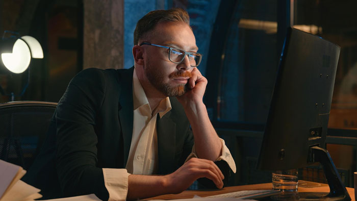 Man in glasses and suit working late at a computer, symbolizing a scammer and hotel receptionist interaction.