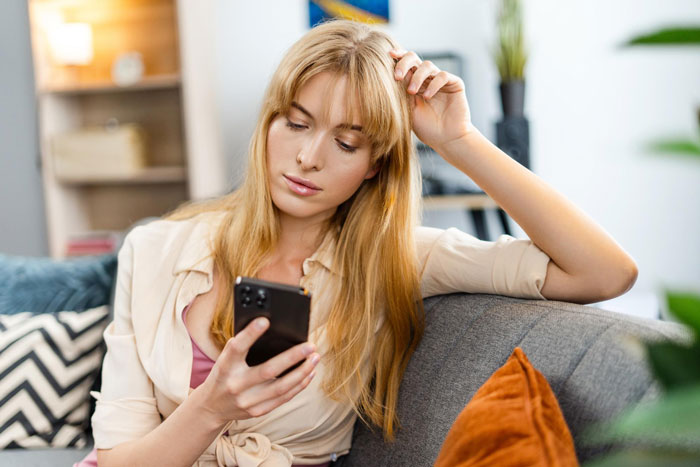 Young woman on couch looking thoughtfully at her phone, reflecting on birthday host drama involving bestie's boyfriend exclusion.