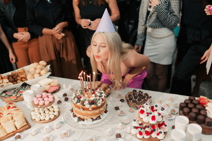 Young woman in a party hat blowing out candles on a birthday cake while guests watch, sparking birthday host drama. Young woman in a party hat blowing out candles on a birthday cake while guests watch, sparking birthday host drama.