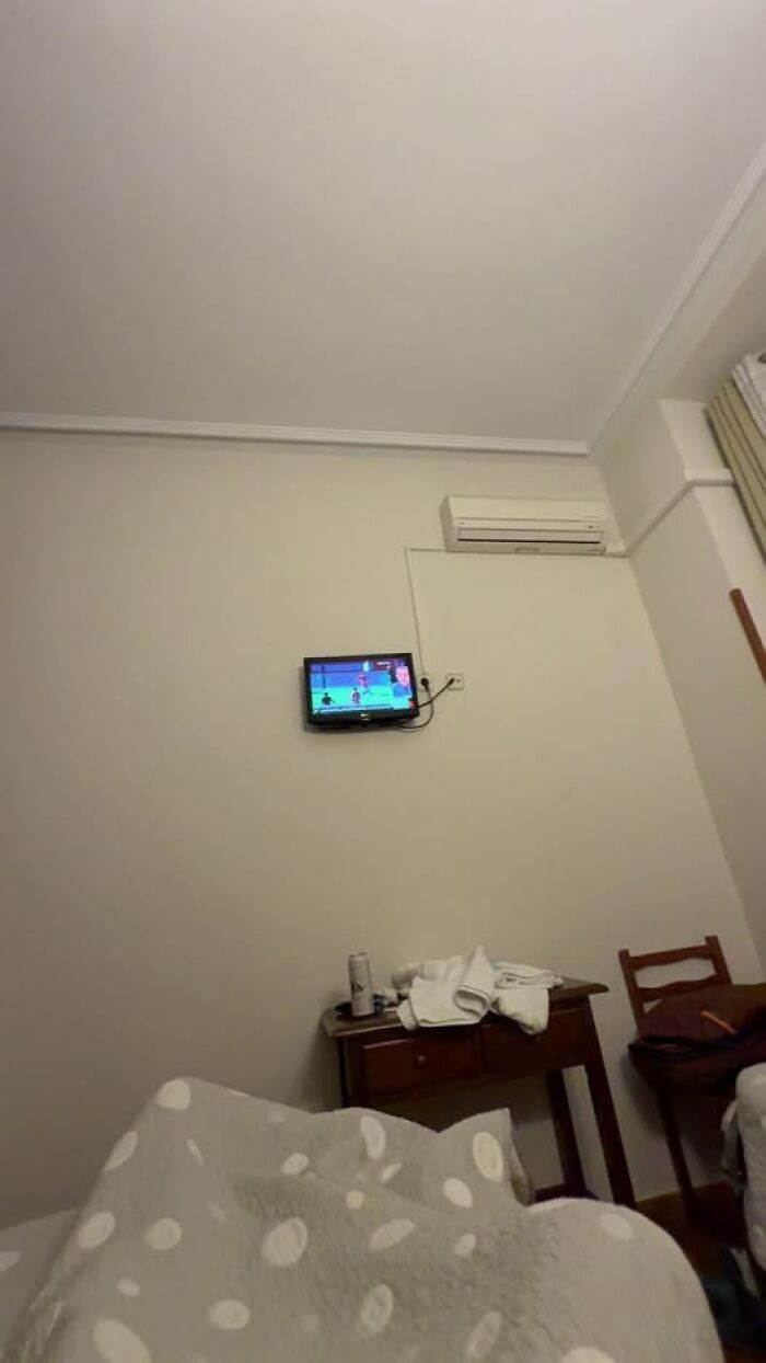 Woke up to small wall TV and air conditioner above bedside table with towels, view from bed in plain hotel room.