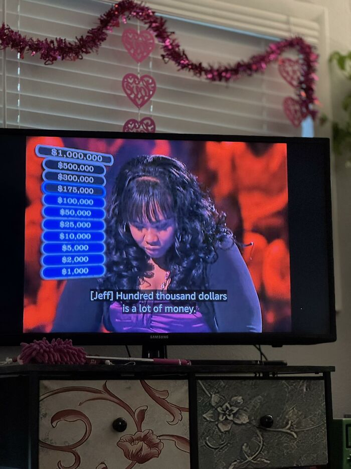 Woman on TV show with disastrous hairstyle and curly bangs, looking down during a high-stakes moment with money prizes displayed.