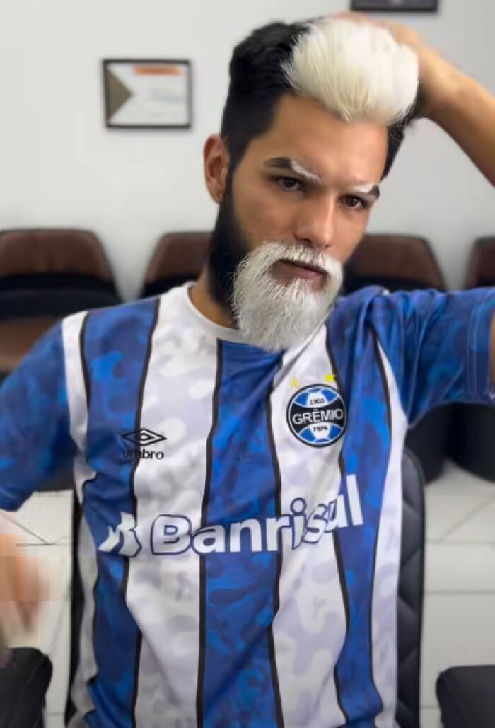 Man with a black and white split dyed hairstyle and beard wearing a blue and white Gremio soccer jersey indoors.
