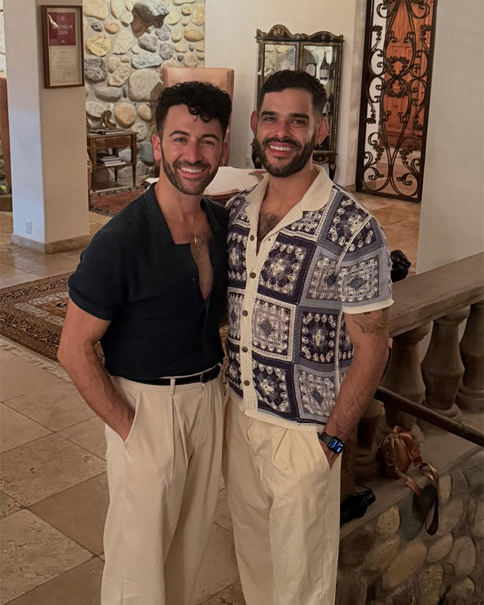 Gay couple smiling and posing indoors, one in patterned short-sleeve shirt and the other in dark open-collar top Gay couple smiling and posing indoors, one in patterned short-sleeve shirt and the other in dark open-collar top