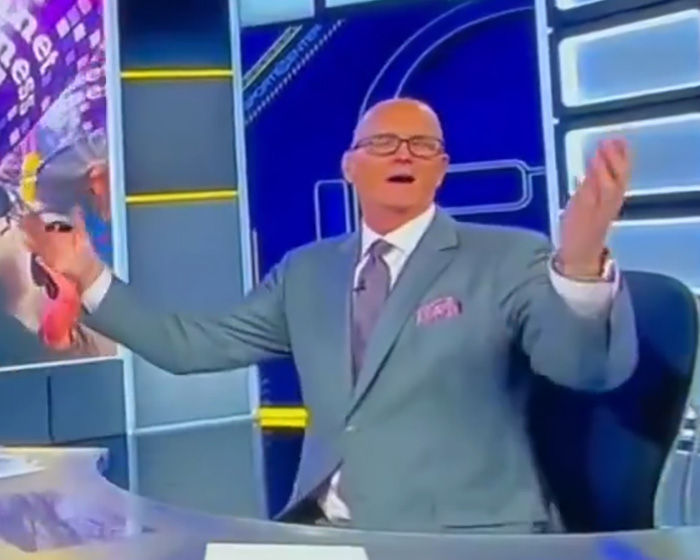 Man in TV studio wearing suit and glasses gesturing at desk, screenshot related to gay couple reaction story Man in TV studio wearing suit and glasses gesturing at desk, screenshot related to gay couple reaction story