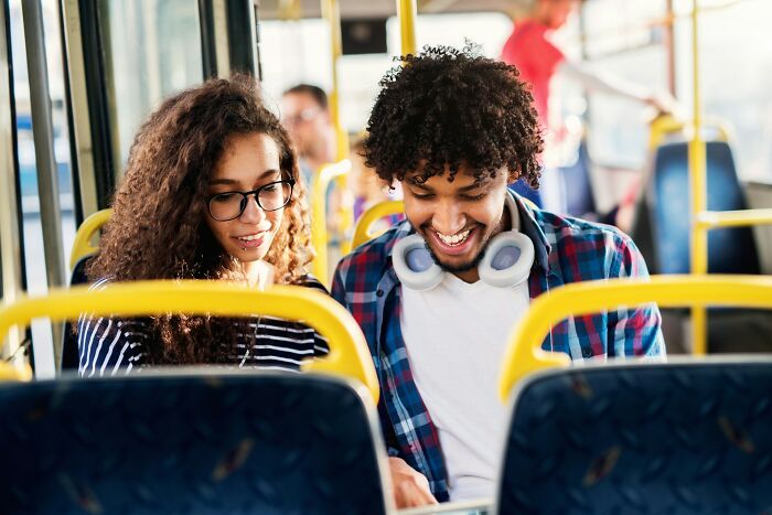 A young man and woman sitting on a bus, smiling and looking at a device, illustrating cultural norms in America.