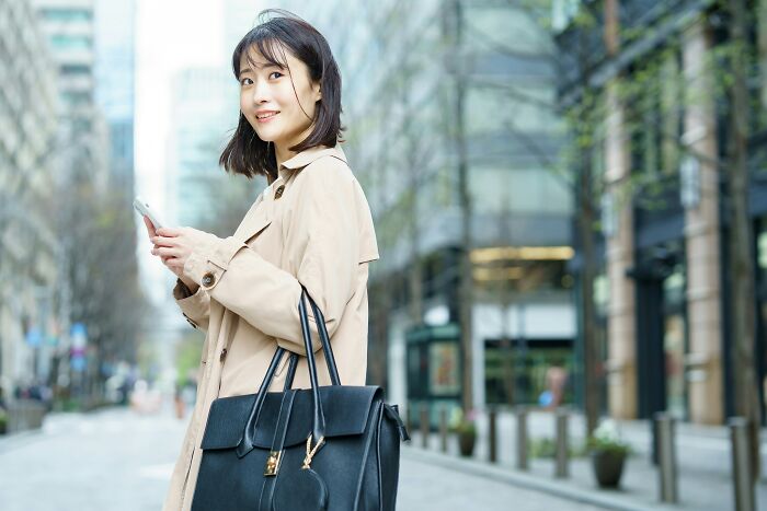 Young woman in a trench coat holding phone and smiling outdoors, representing coworkers acting unhinged stories.