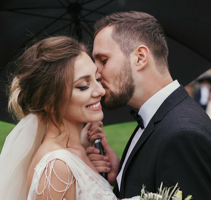 Bride smiling as groom kisses her on the cheek under umbrella, capturing wedding moments that turned out to be accurate