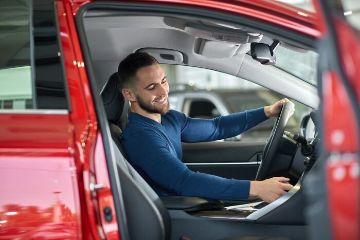 Man with short hair sitting inside a red car, smiling while checking the dashboard, illustrating overrated adult goals.
